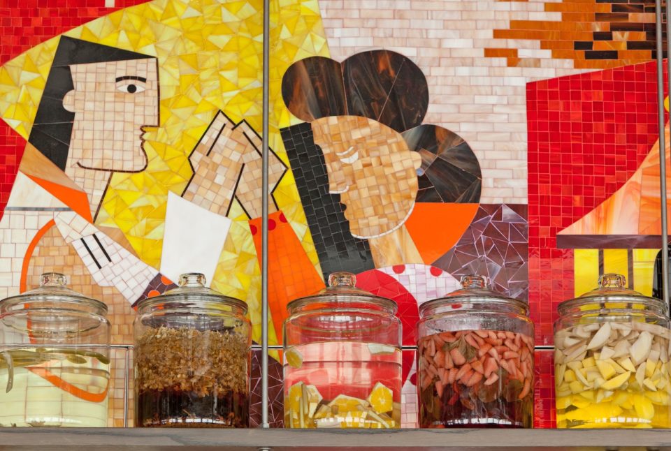 Glass jars of tequila with various fruit infusions sit on a shelf in front of a mosaic wall depicting cubist art.