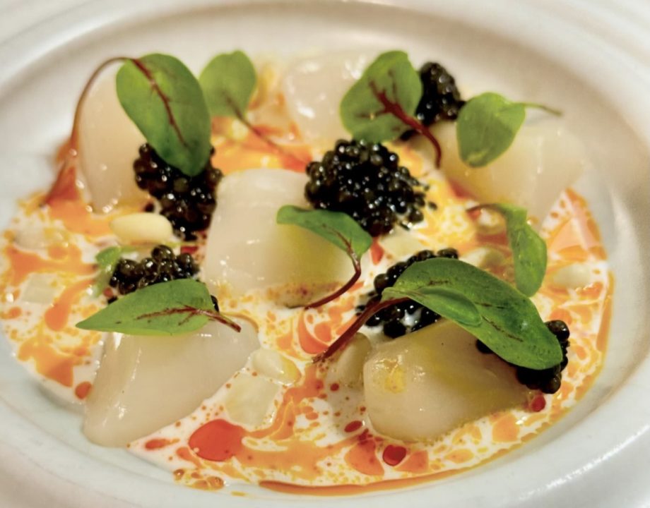 Closeup of a shallow white bowl holding six raw scallops, several small spoonfuls of caviar, and green herbs, in a red and while swirled sauce.