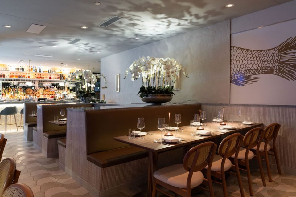 A dimly lit restaurant dining room with three dark wood booths, padded wooden chairs, large orchid arrangments on shelves between the tables, and a wall painting of a fish's tail.
