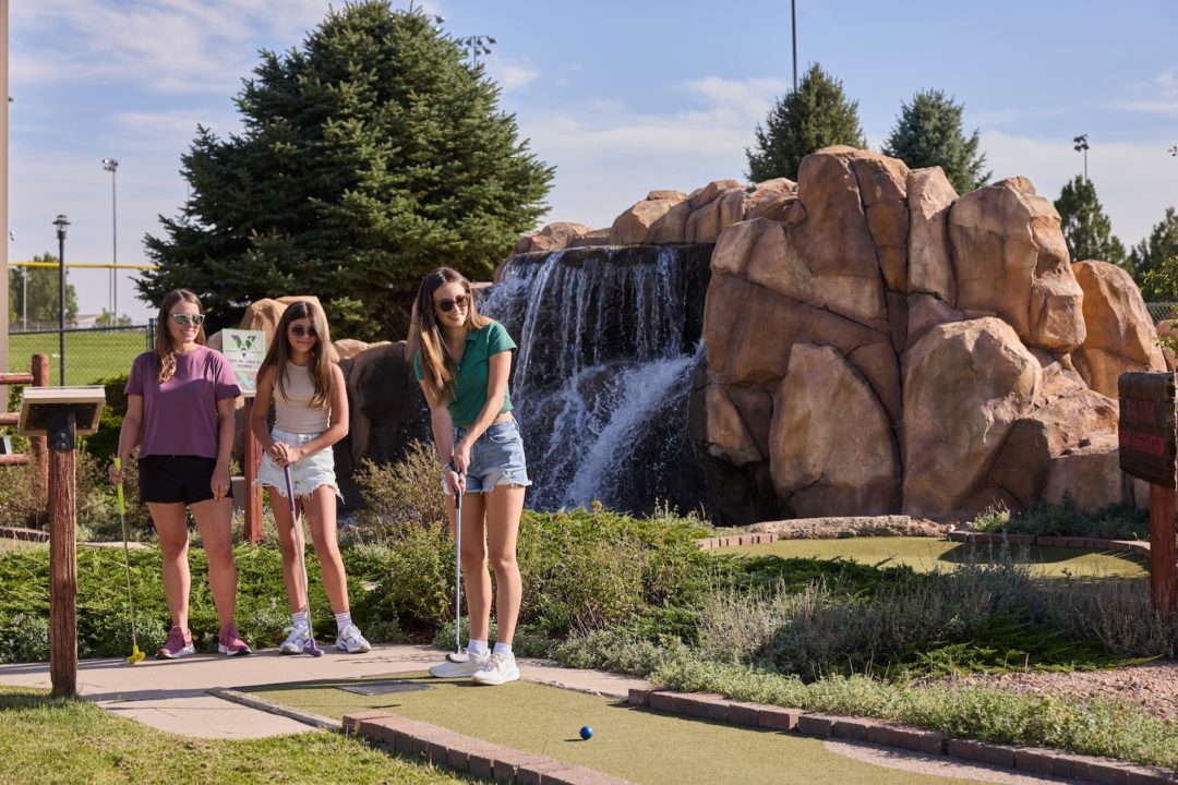 Three girls play mini golf in front of an artificial waterfall at the Greeley Family Fun Plex