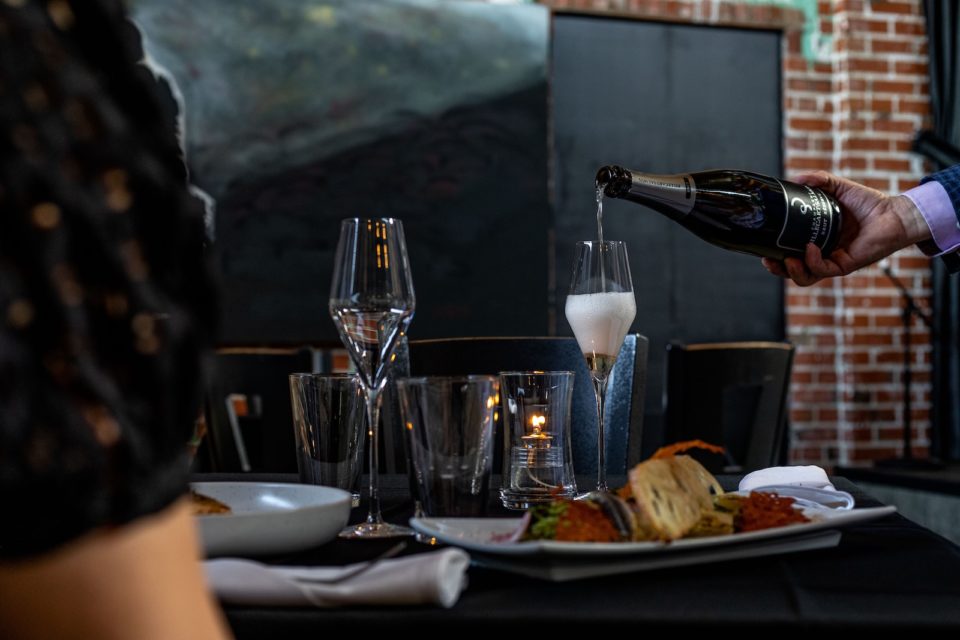 A waiter's hand pouring champagne into a tall glass at a dinner table with a plate of food.