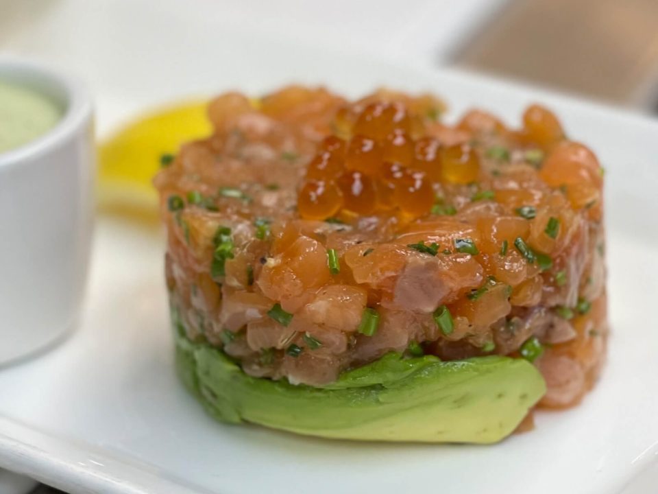 Dice raw salmon and salmon roe on top of slices of avocato on a white plate with lemon wedge and a ramekin of green sauce in the background.