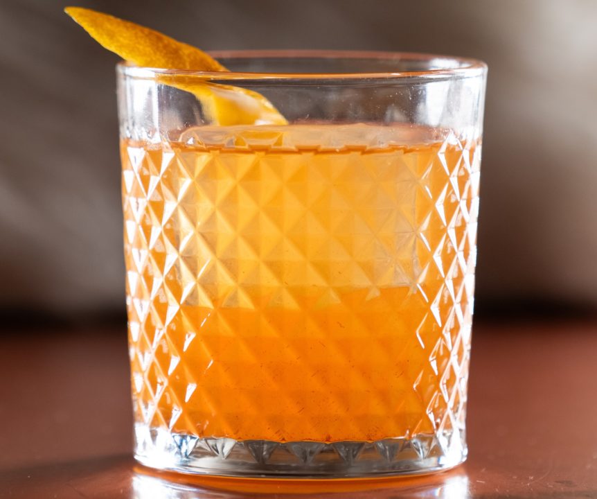 Closeup of a bright orange cocktail in a rocks glass with a lemon peel garnish.