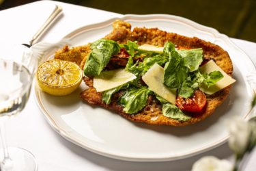 A breaded veal cutlet topped with lettuces, parmesan, tomatoes and half of a grilled lemon
