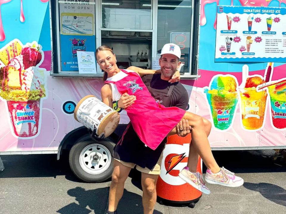 A man in a black apron is holding his wife in a pink apron, whi is holding a large ice cream container in front of an ice cream truck