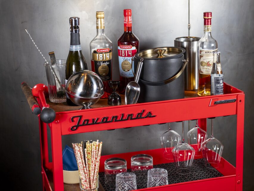 A red bar cart loaded with bottles of Italian spirits and glassware.