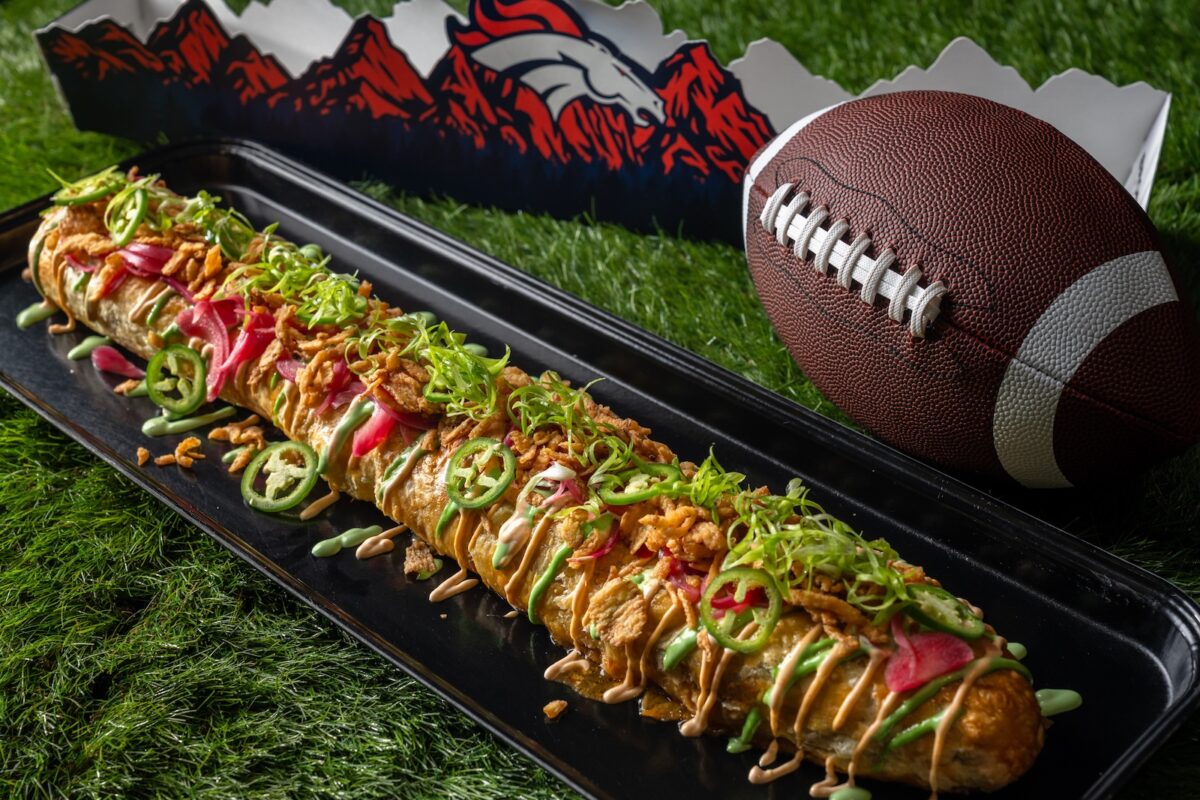A two-foot hot dog wrapped in puff pastry with typical hot dog toppings next to a football and a Denver Broncos pendant