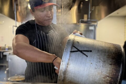A Hispanic man in a kitchen scoops food from a large pot.