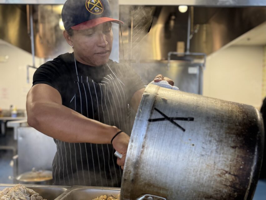 A Hispanic man in a kitchen scoops food from a large pot.
