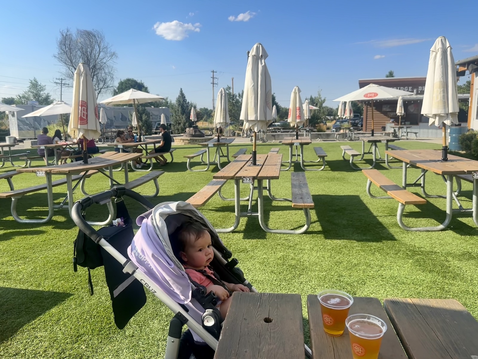 A child in a stroller on a turf-covered restaurant patio with picnic tables and closed umbrellas