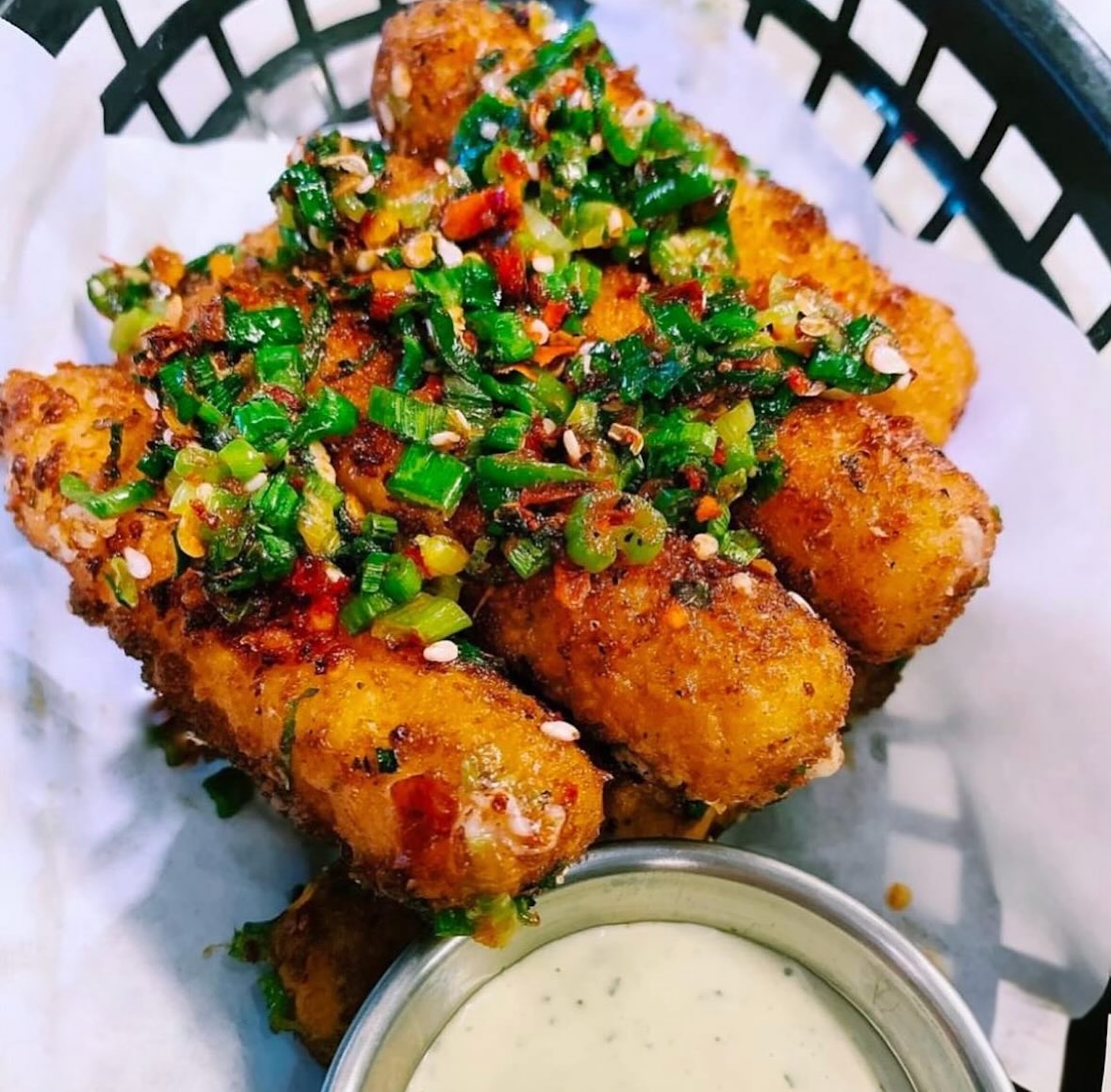 Several fried mozzarella sticks topped with fresh herbs and chiles with creamy dipping sauce in a paper-lined black plastic basket
