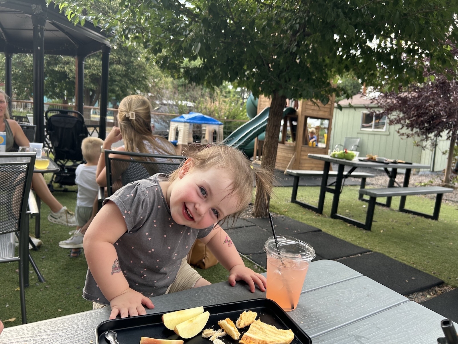 Child with tray of food and pink drink at a restaurant patio table with playground in the background.