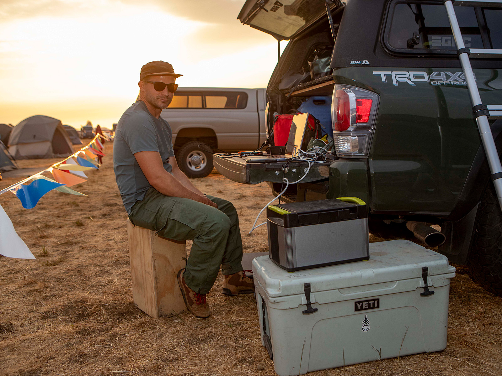 Man sitting on a box behind a car working on his computer