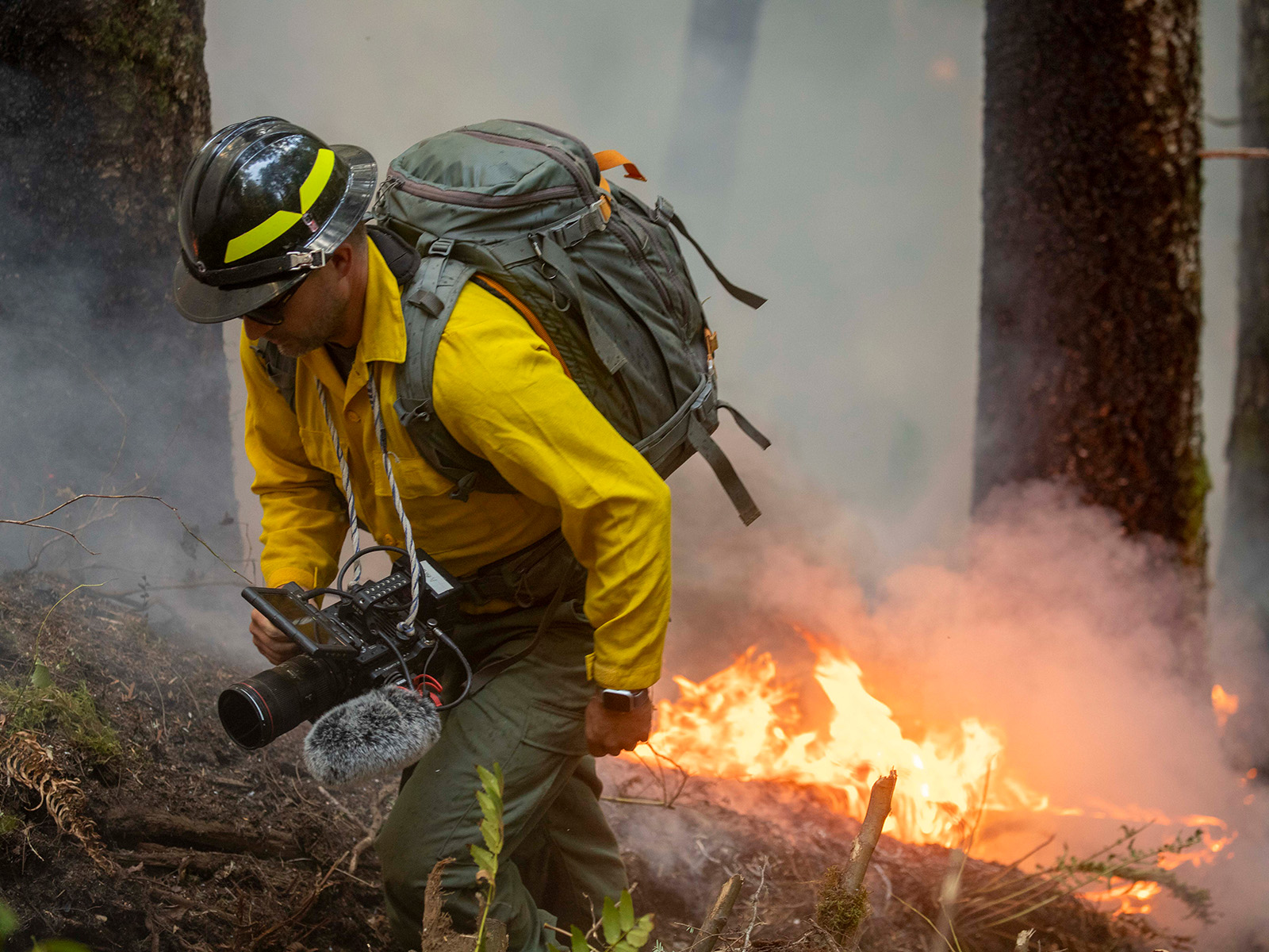 Man holding a camera dressed in firefighting career walks away from a fire in the forest