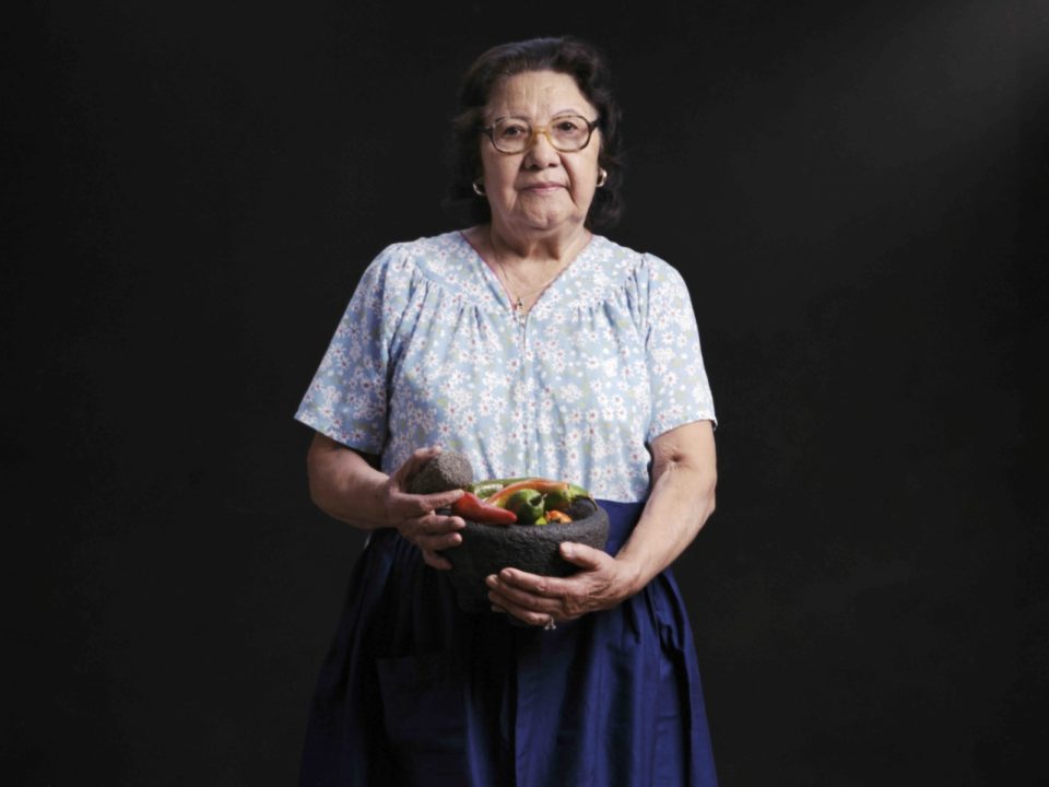 A Mexican woman wearing a skirt and blouse holds a stone mortar and pestle with chiles.