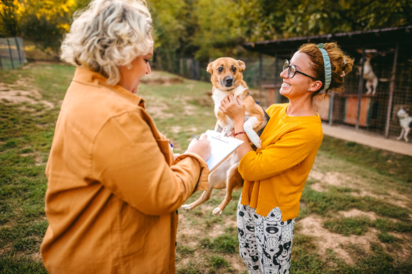 Two women standing outside. One holds a dog, while the other takes notes.