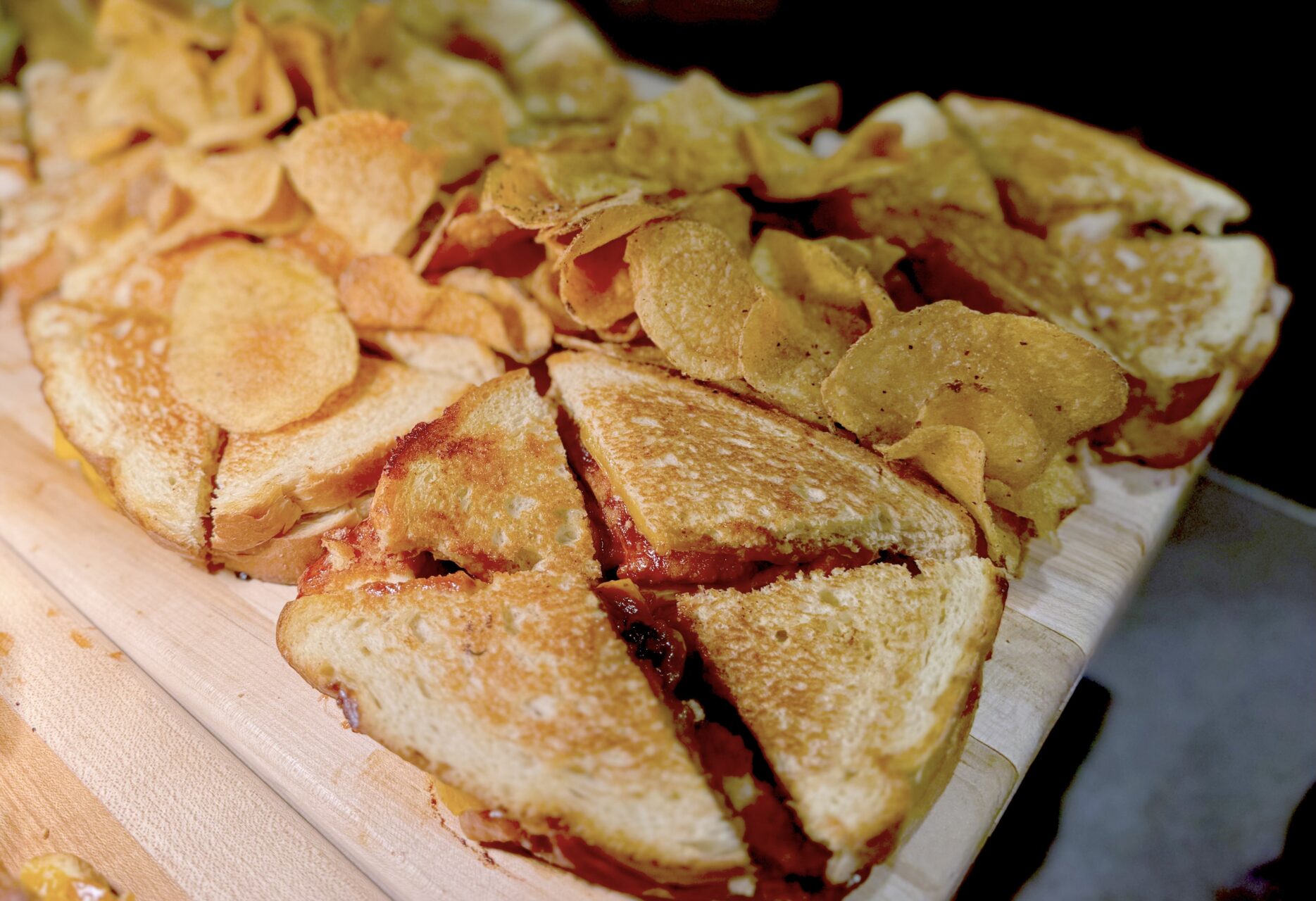 A wooden board with grilled sandwiches cut into quarters and a pile of potato chips