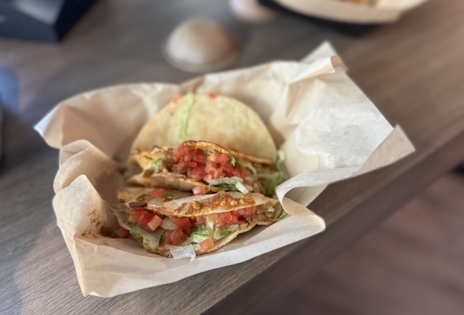 A trio of tacos topped with diced tomatoes and lettuce in a paper-lined basket