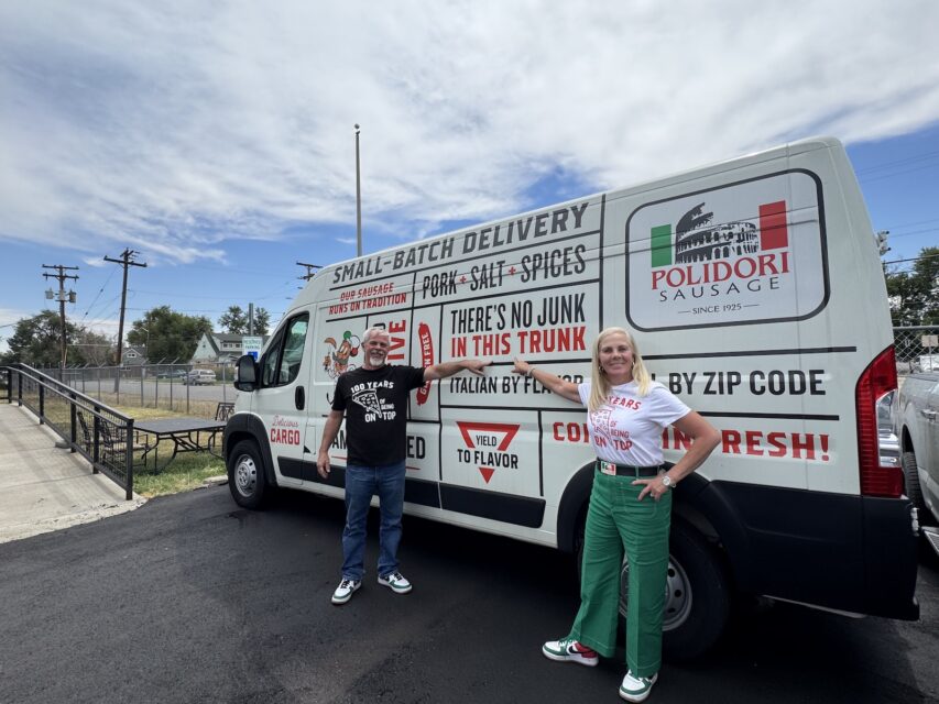 A man in a black T-shirt and a woman in a white T-Shirt and green pants stand in front of a Polidori Sausage van.