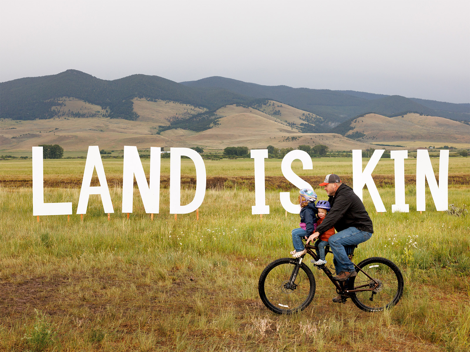 A man bikes past a sign that says Land Is Kin; he has two children on the bike with him