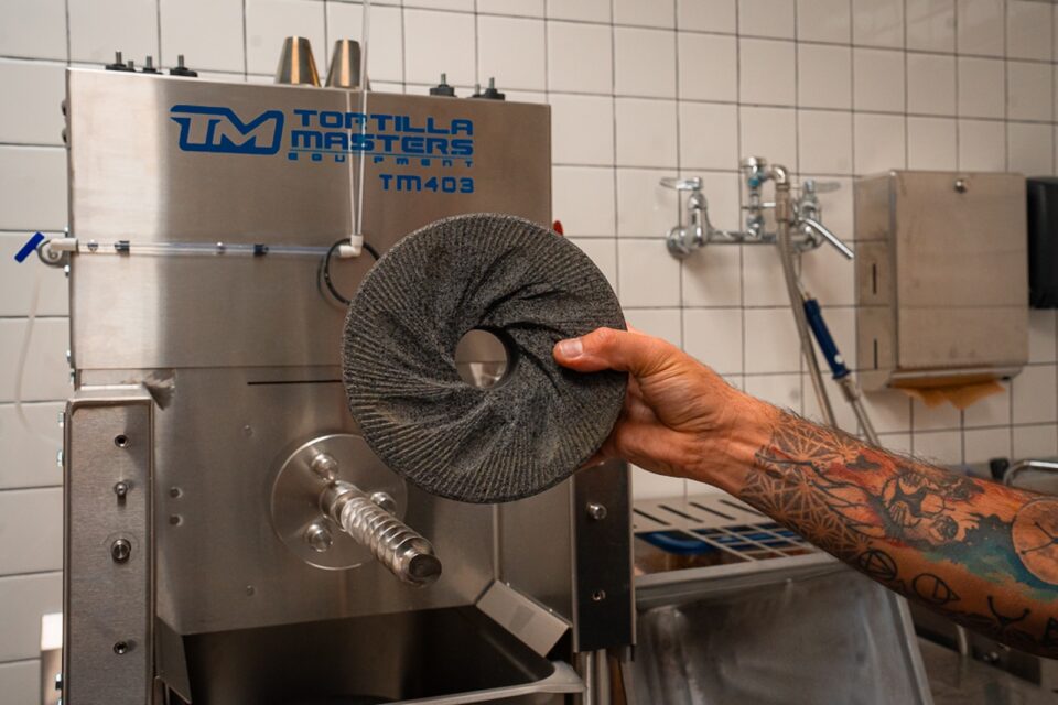 A tattooed arm with hand holding a stone grinding wheel in front of a corn grinding machine