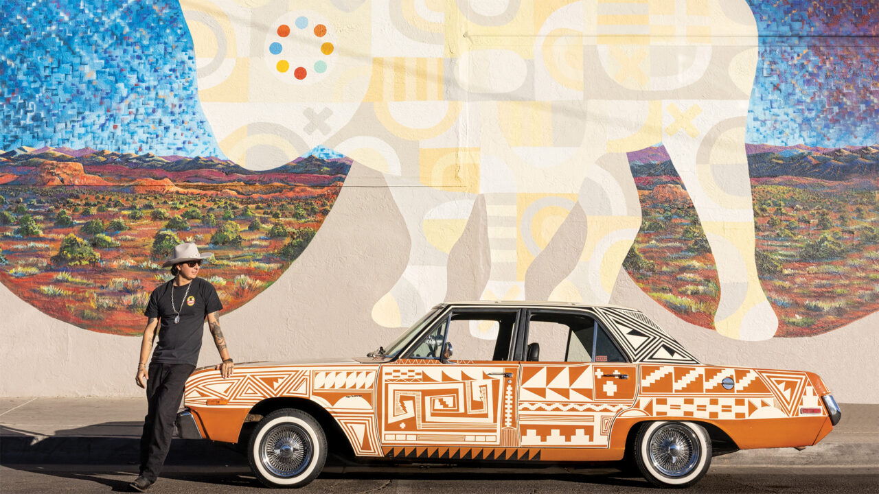 An artist stands next to an orange car in front of a mural