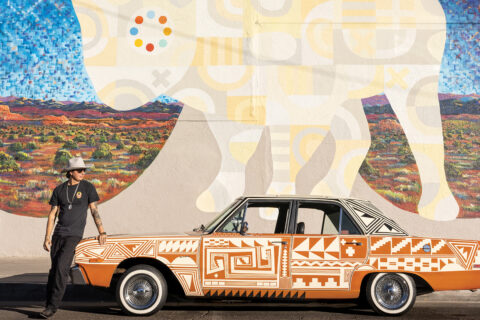 An artist stands next to an orange car in front of a mural