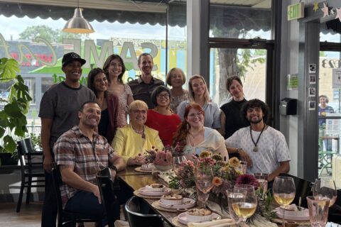 A group of 11 people sit and stand at one end of a set restaurant table with chef Lidia Bastianich in the center