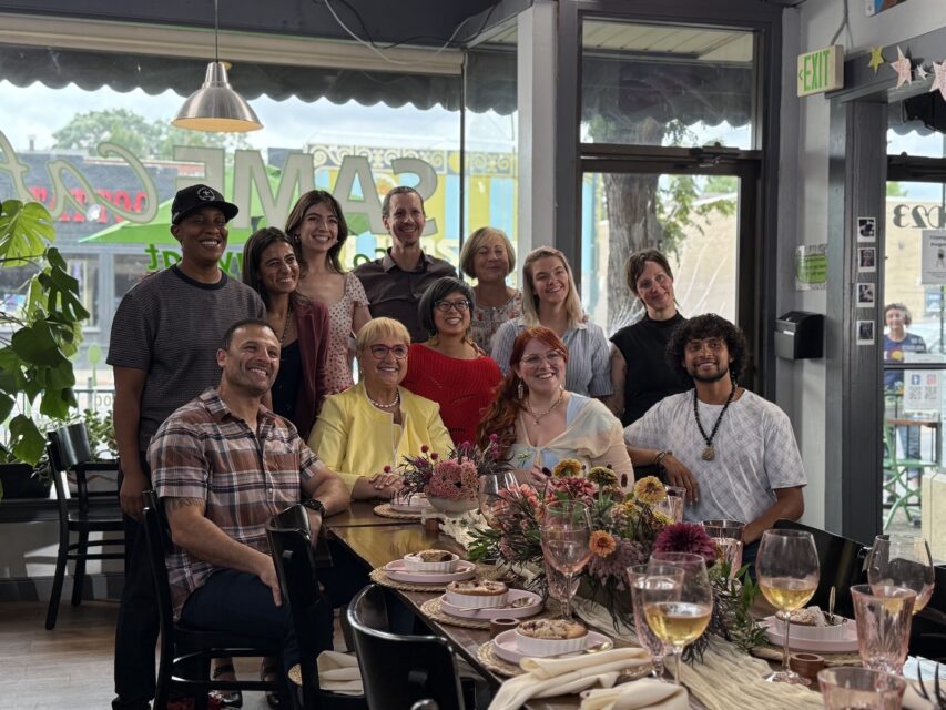 A group of 11 people sit and stand at one end of a set restaurant table with chef Lidia Bastianich in the center