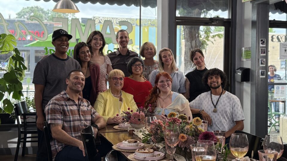 A group of 11 people sit and stand at one end of a set restaurant table with chef Lidia Bastianich in the center