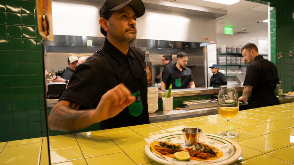A chef in a black baseball hat stands between the kitchen and a yellow tile countertop with a plate of two tacos