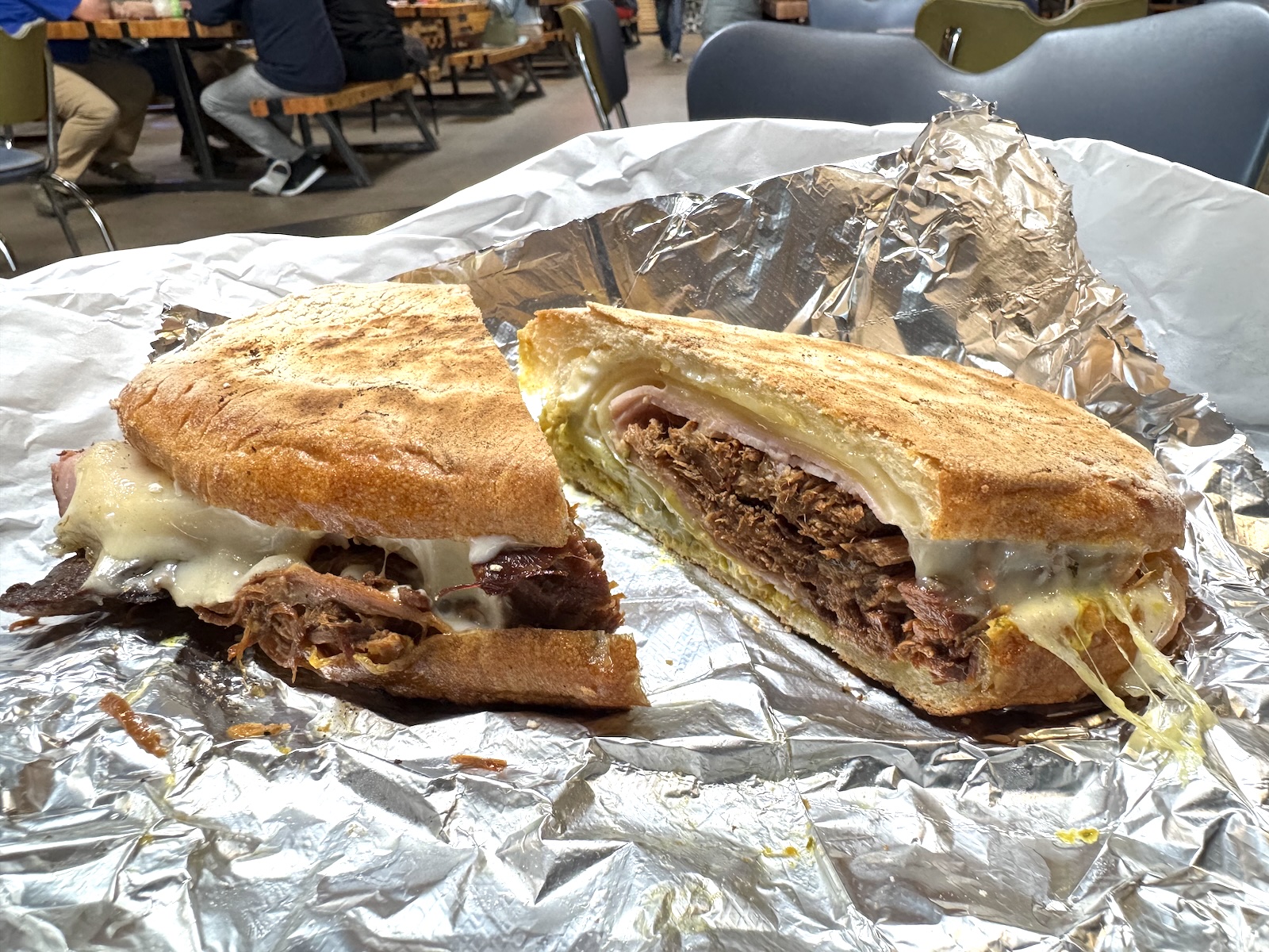 A grill-pressed Cuban sandwich cut in half and filled with shredded pork, ham, Swiss cheese, pickles, and mustard on an open foil wrapper