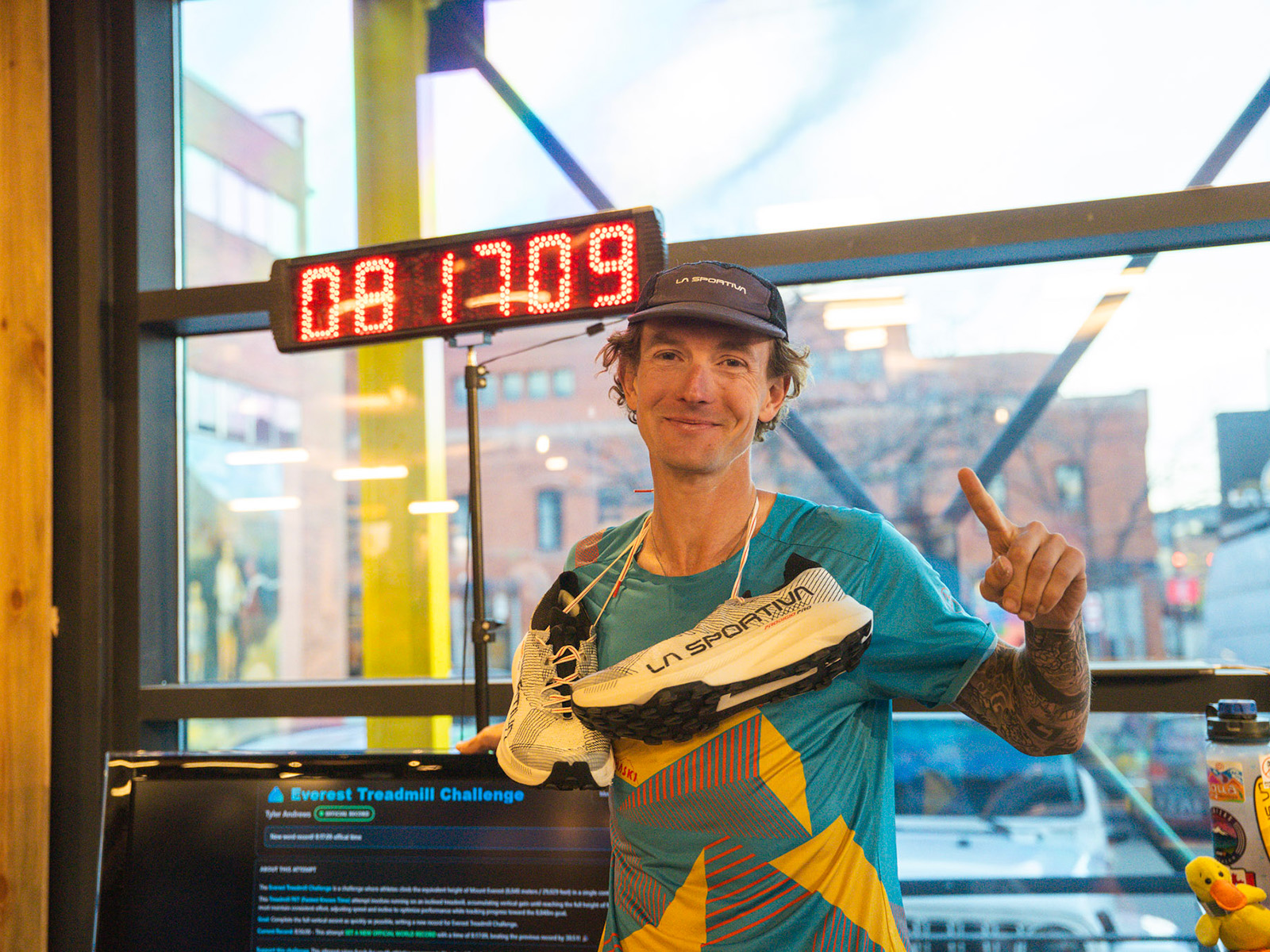 Tyler Andrews poses in front of a treadmill with one finger up and sneakers around his neck