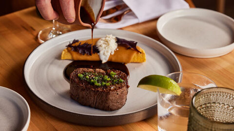 A steak on a white plate with a hand pouring a gravy over it.