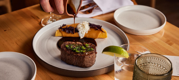 A steak on a white plate with a hand pouring a gravy over it.