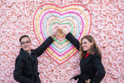 Two people standing together in the middle of a heart, who have their hands placed together in a heart shape.