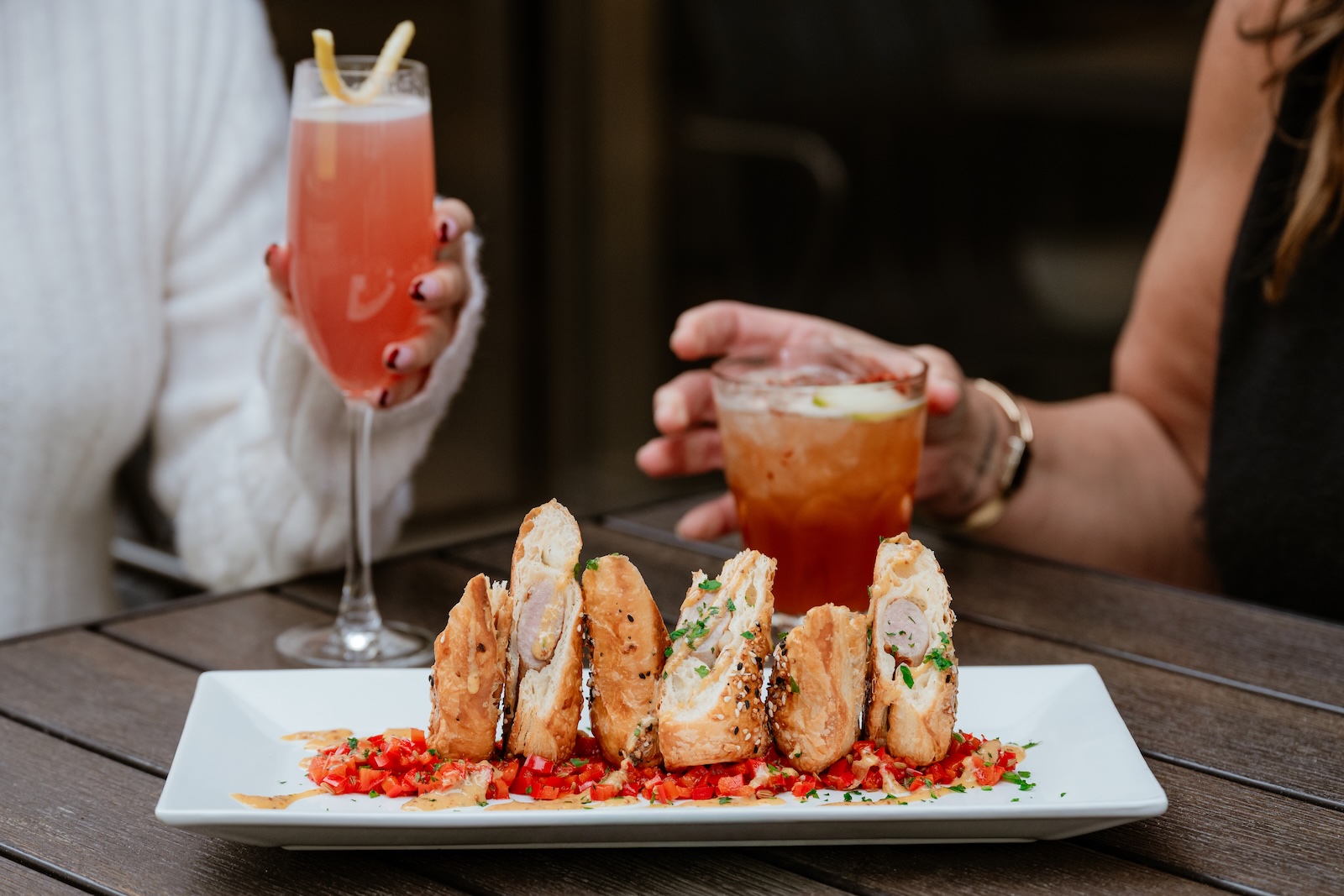 A plate of sausage and puff pastry rolls sliced diagonally and stood up vertically in red sauce on a rectangular white plate with two hands holding cocktails in the background
