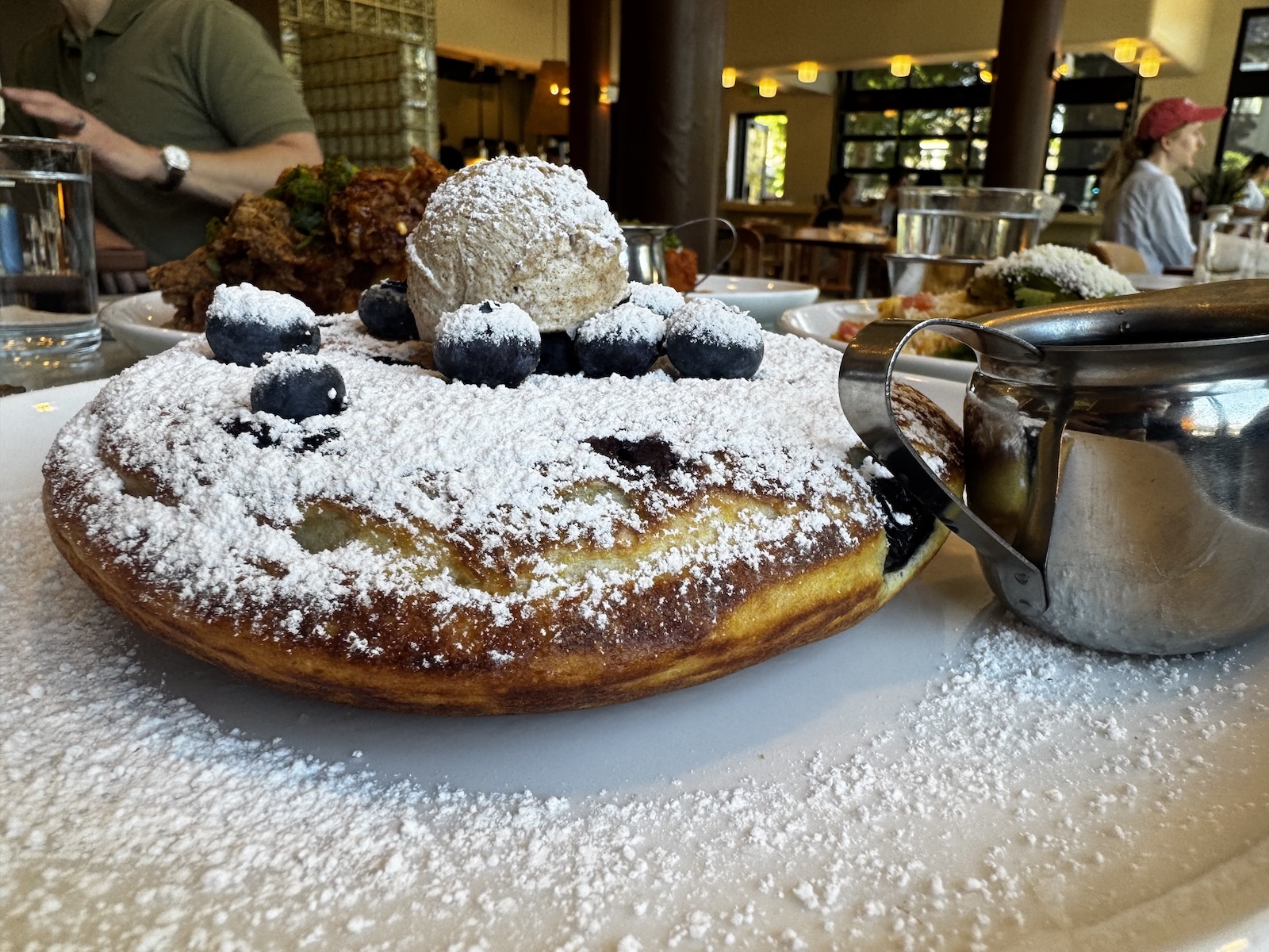 A single large pancake topped with powdered sugar, a scoop of butter, and blueberries with a small silver syrup pitcher.