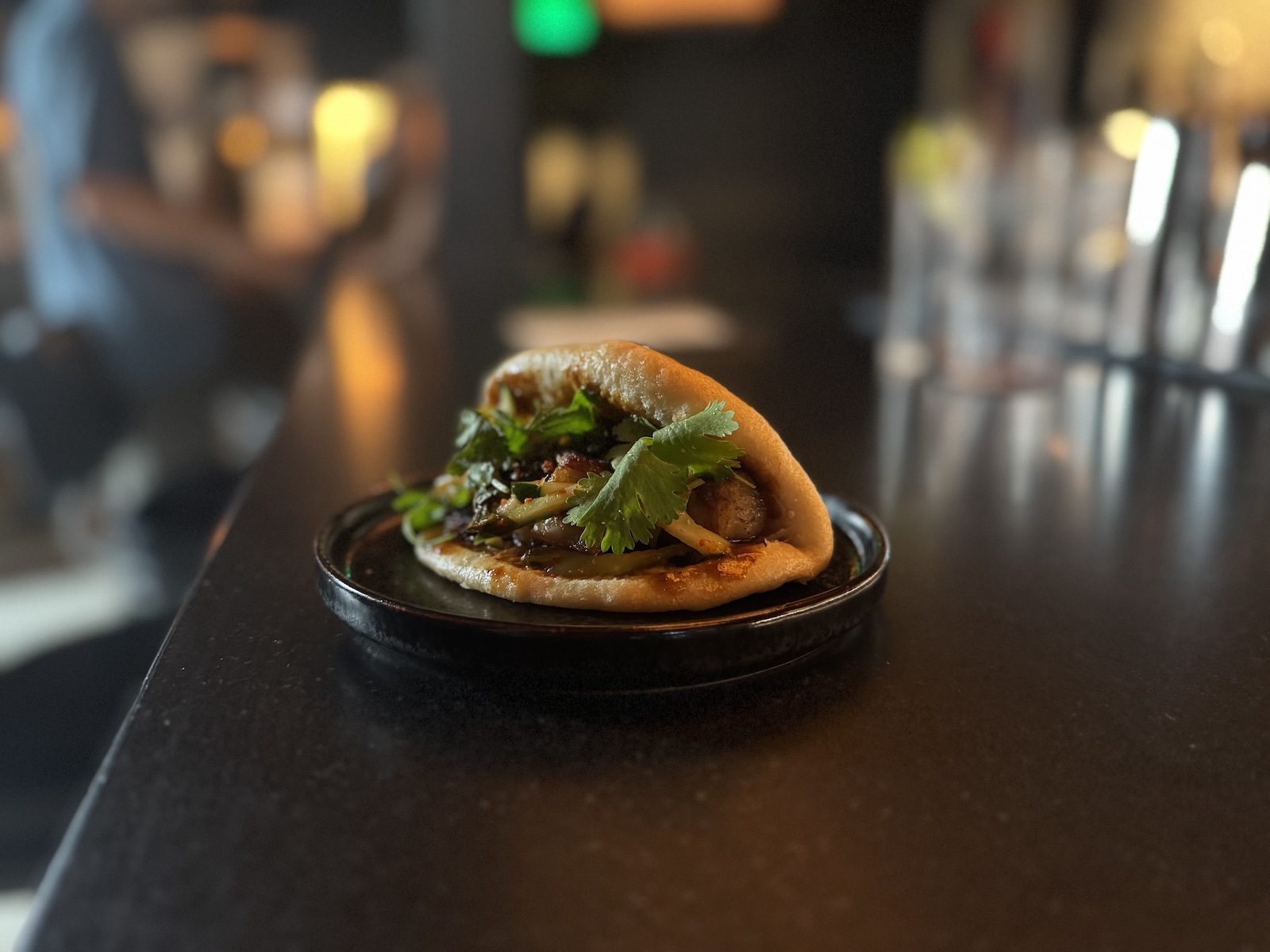 A single Japanese bao bun stuffed with pork belly and greens on a black plate.