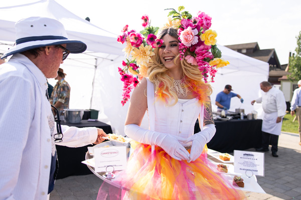 A woman standing outside with pink and yellow flowers in her hair like a giant crown, while wearing a white shirt and pink and yellow skirt.