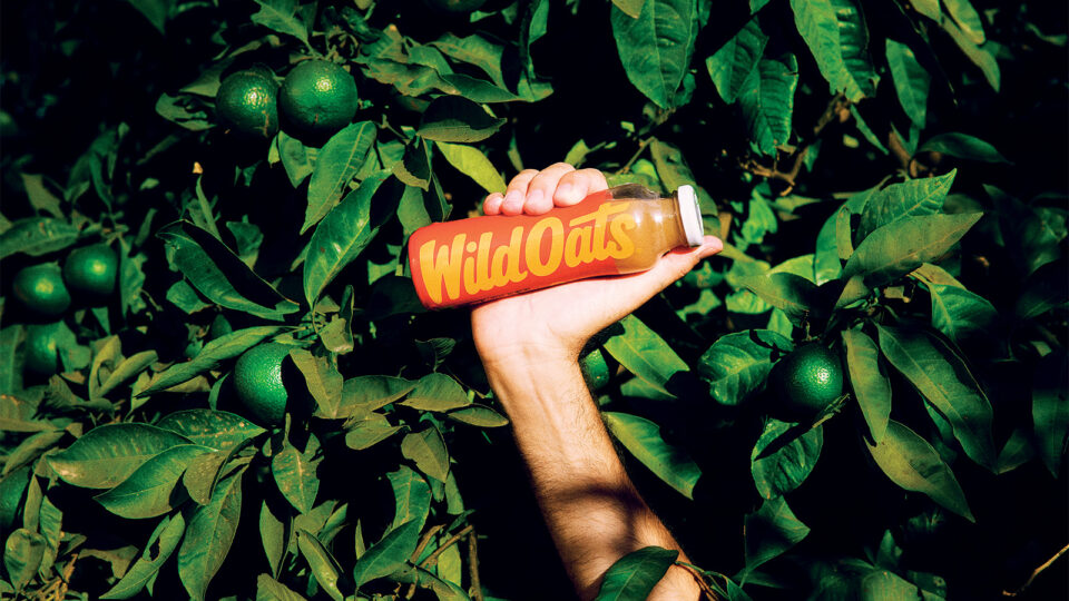 A hand holding Wild Oats juice, sticking out from a hedge