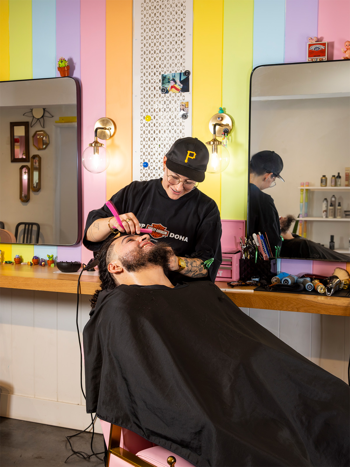 A barber wearing a Pittsburgh Pirates hat works on a man with a beard in a colorful salon