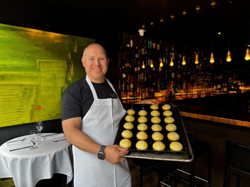 A pastry chef in a white apron holds a silicone tray with 20 round dough balls rising
