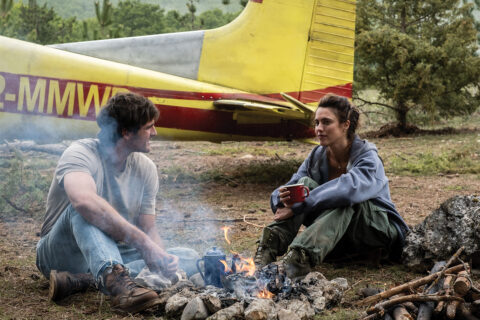 A man and a woman sitting in the dirt by a campfire in front of a yellow Cessna
