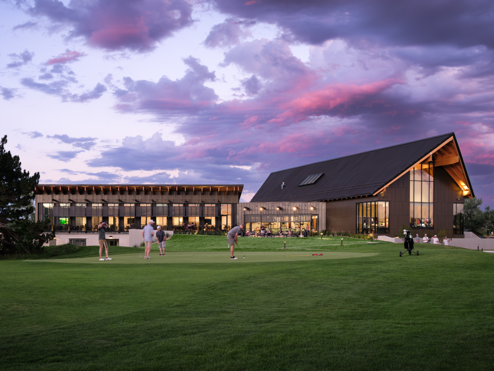 Golfers practice putting on a green outside a contemporary golf clubhouse with expansive glass windows, set against a stunning sunset sky.