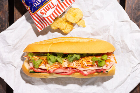 A banh mi on white paper with a bag of potato ships
