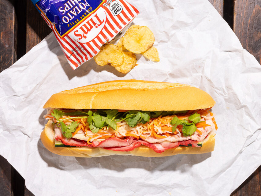 A banh mi on white paper with a bag of potato ships