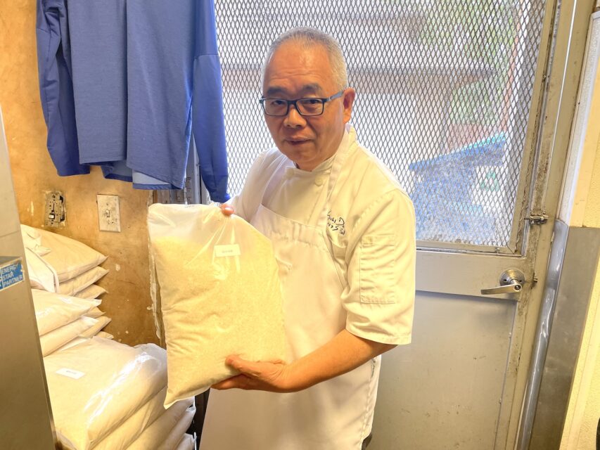 A Japanese man in a chef coat holding a large plastic bag of rice