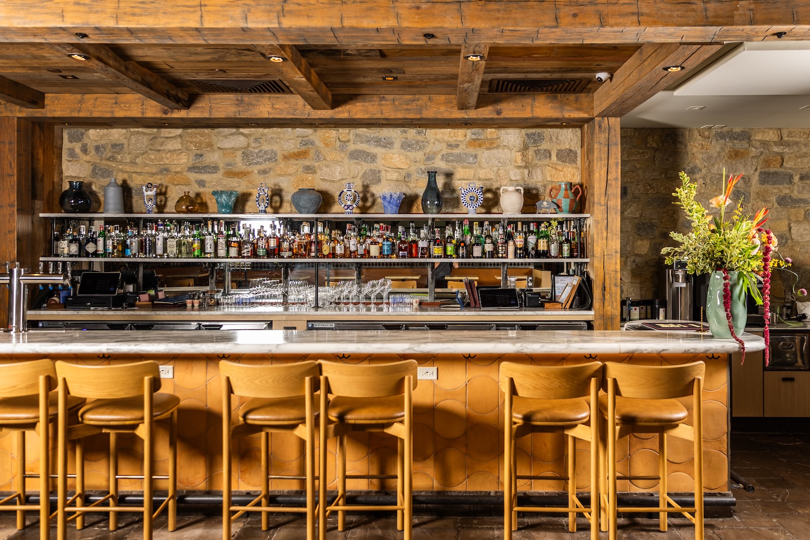 A bar with several bar stools, shelves of liquor bottles, a cobblestone wall behind the shelves, and heavy wood beams above the bar.