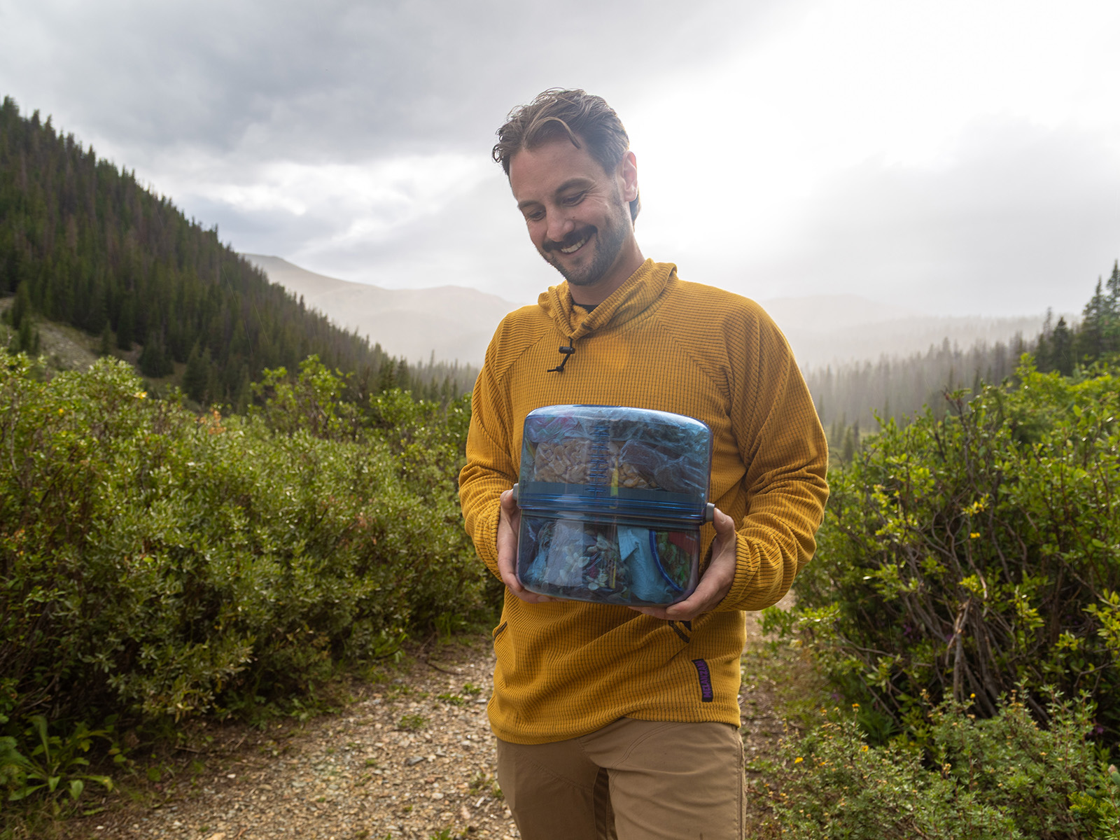 Man holds clear blue bear canister filled with food in the mountains